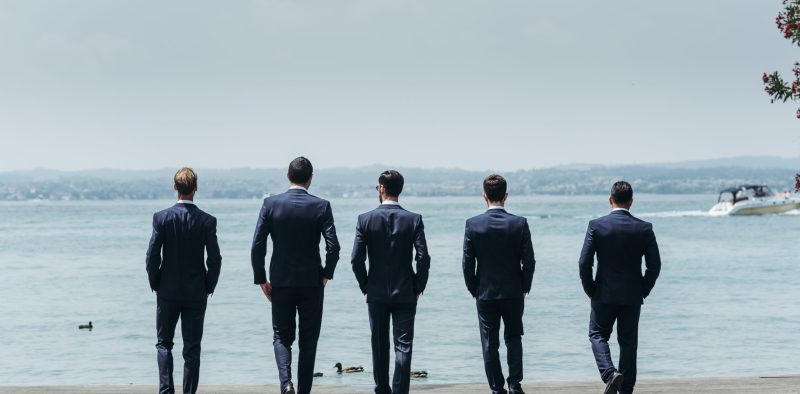 Five men in classy suits walk towards the blue sea